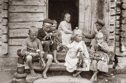 Mustavalkoinen valokuva, jossa kuusi lasta istuu hirsirakennuksen portailla syömässä tuoreita nauriita. Taustalla aikuinen nainen.