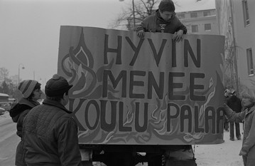 Poika pitelemässä maalattua kylttiä "Hyvin menee koulu palaa". 1970-luku. Kuva: Lauri Pietarinen. Helsingin kaupunginmuseo. CC BY 4.0.