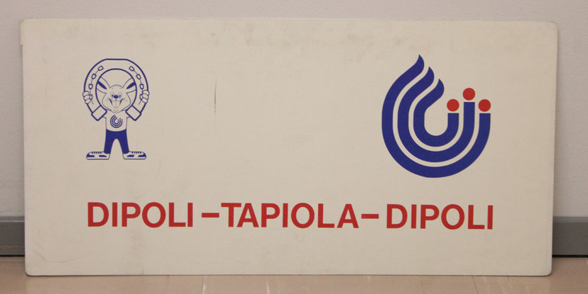 Dipoli–Tapiola–Dipoli. Valkoinen muovikyltti, jossa kuva-aiheena yleisurheilun MM-kisojen 1983 logo ja maskotti Lasse-jänis, joka nostaa hevosenkenkää. Kuva: Urheilumuseo. CC BY-ND 4.0.