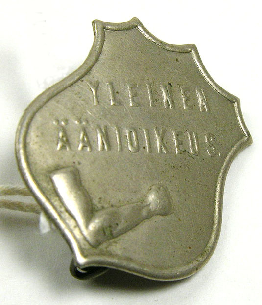 Yleinen äänioikeus -rintamerkki. Vaakunakilven muotoisessa merkissä koukkuun taivutettu käsivarsi nyrkissä. Kuva: Turun museokeskus. CC BY-ND 4.0.