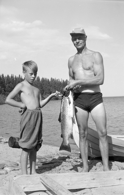 Lauri Mikkonen kädessään Oulujärvestä pyydetty kuuden kilon painoinen järvitaimen. Vieressä Mikkosen poika. 1958. Kuva: Museovirasto. CC BY 4.0.
