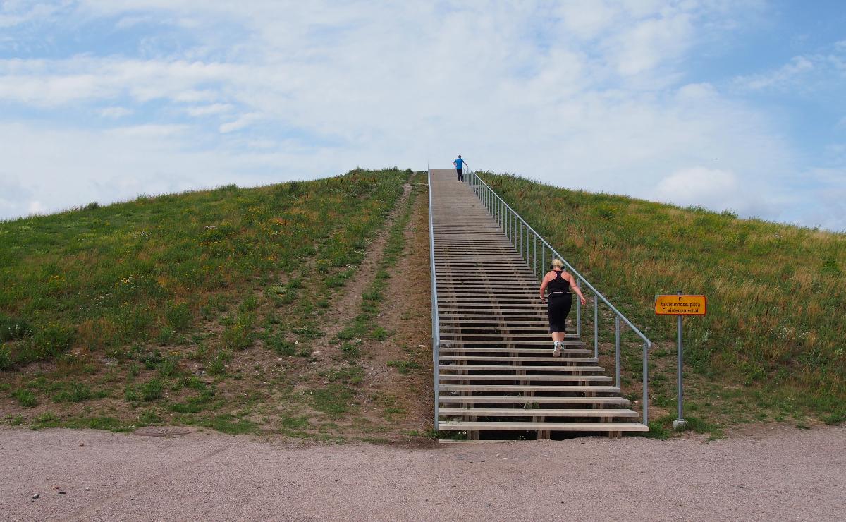 Ruohon peittämä mäki, jonka huipulle nousee pitkät ja leveät puuportaat. Portaissa kaksi juoksijaa.