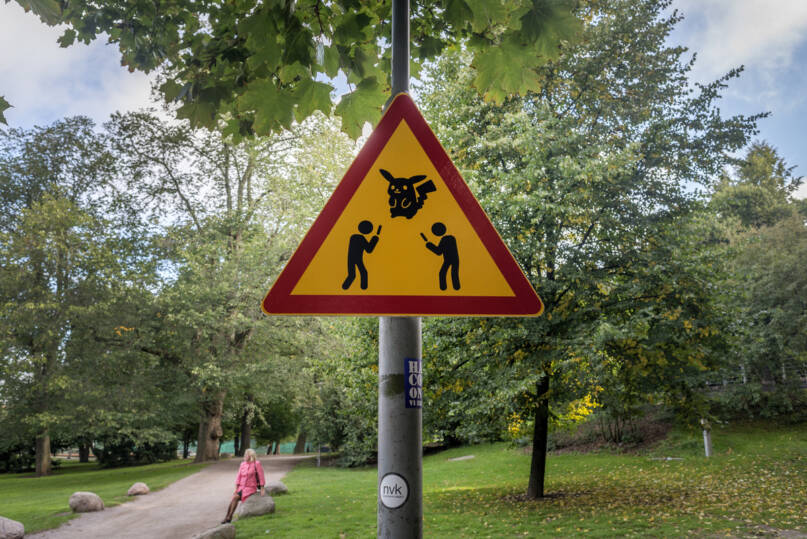 Puistomaisemaa ja varoituskolmio, jossa on Pokemon-eläinhahmo sekä kaksi ihmishahmoa kävelemässä puhelin kädessä.