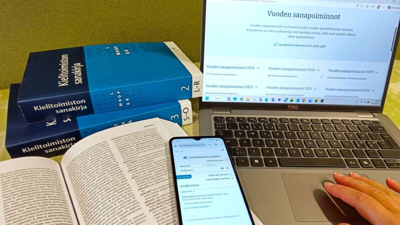 Asetelma, jossa taustalla kaksi Kielitoimiston sanakirjan painettua osaa sekä kannettava tietokone, jonka näytöltä näkyy Vuoden sanapoimintoja -julkaisu. Etualalla avattu sanakirja, jonka päällä on puhelin. Puhelimen näytölle on avattu uudissana-artikkeli sähköisestä Kielitoimiston sanakirjasta.