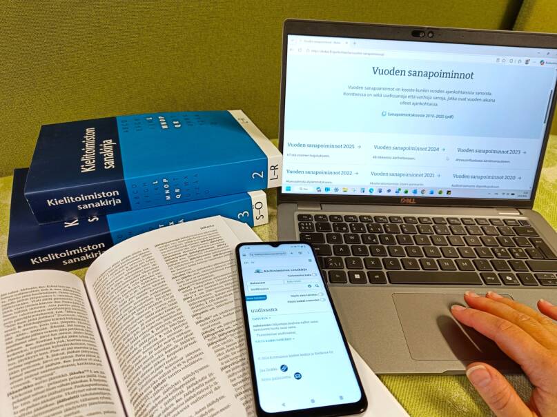 Asetelma, jossa taustalla kaksi Kielitoimiston sanakirjan painettua osaa sekä kannettava tietokone, jonka näytöltä näkyy Vuoden sanapoimintoja -julkaisu. Etualalla avattu sanakirja, jonka päällä on puhelin. Puhelimen näytölle on avattu uudissana-artikkeli sähköisestä Kielitoimiston sanakirjasta.