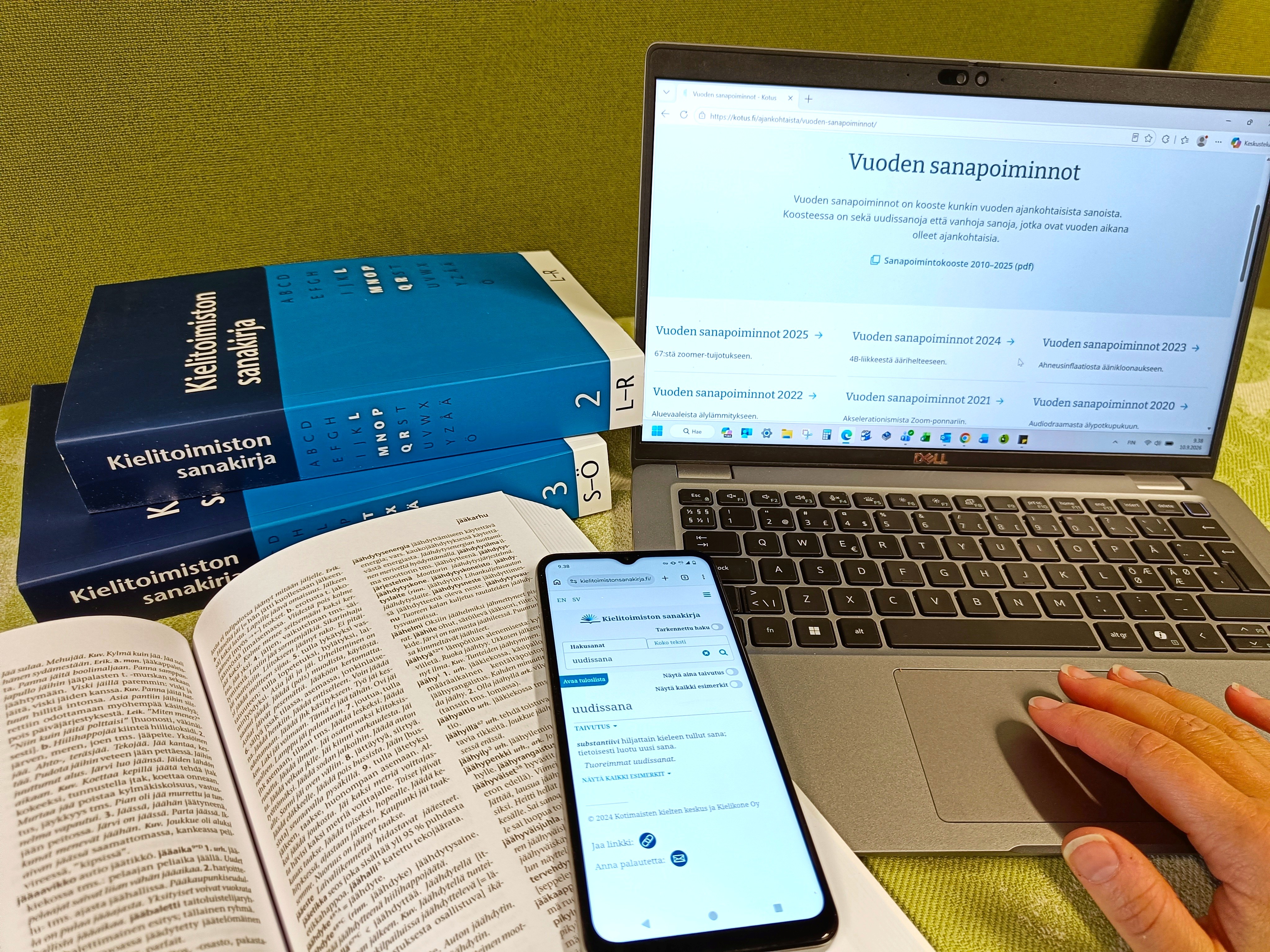 Asetelma, jossa taustalla kaksi Kielitoimiston sanakirjan painettua osaa sekä kannettava tietokone, jonka näytöltä näkyy Vuoden sanapoimintoja -julkaisu. Etualalla avattu sanakirja, jonka päällä on puhelin. Puhelimen näytölle on avattu uudissana-artikkeli sähköisestä Kielitoimiston sanakirjasta.