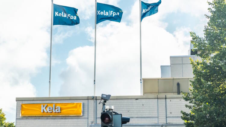 Kelan toimipiste kuvattuna ulkoa. Etualalla koivu sekä jalankulkijoille tarkoitettu liikennevalo, jossa palaa vihreä valo.